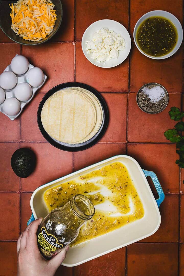 Hand pours jar of green salsa into baking dish for a huevos rancheros breakfast casserole, surrounded by eggs, cheese, tortillas, and seasonings.