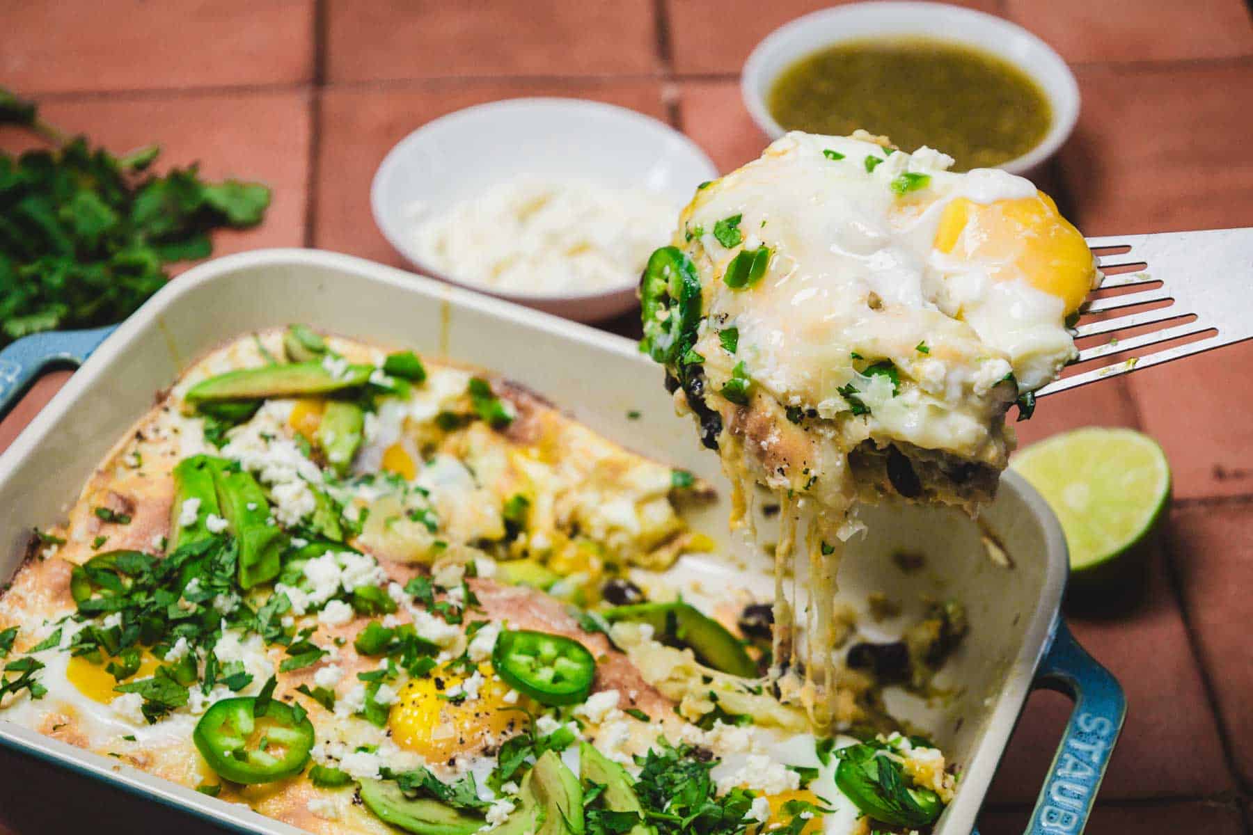 A serving of cheesy baked eggs with jalape&ntilde;os, herbs, and sauce is lifted from a huevos rancheros breakfast casserole.