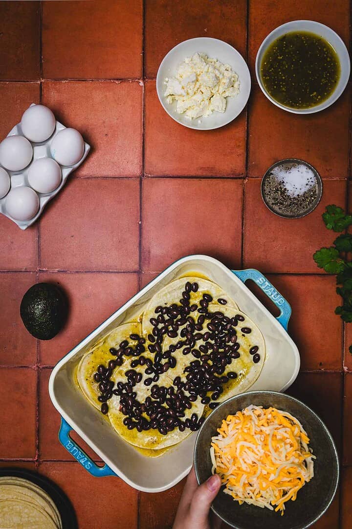 Huevos rancheros breakfast casserole served in a baking dish with beans, shredded cheese, eggs, feta, savory sauce, salt, pepper, and creamy avocado on a red tile counter.