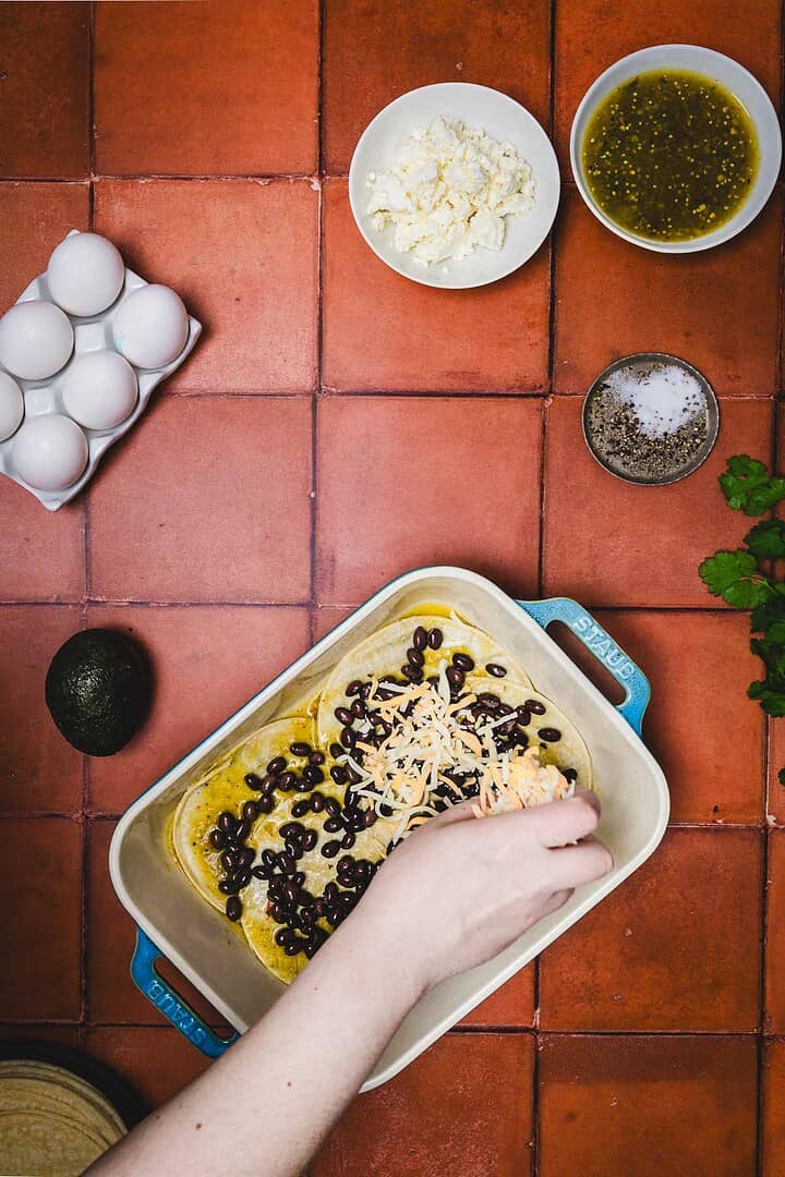 A hand adds cheese to a baking dish with beans for a delicious huevos rancheros breakfast casserole, surrounded by eggs, avocado, cheese, and seasonings.