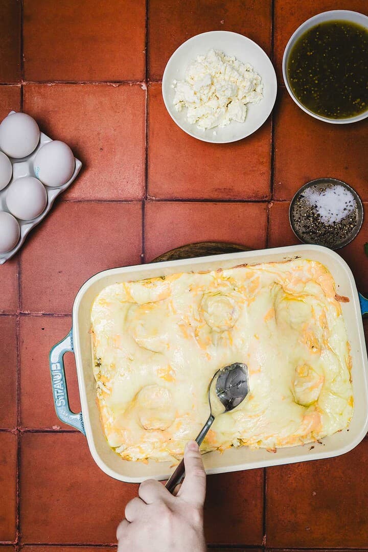 Hand scooping cheesy huevos rancheros breakfast casserole in a blue dish; eggs, feta, herbs, salt, and pepper on red tiled counter.