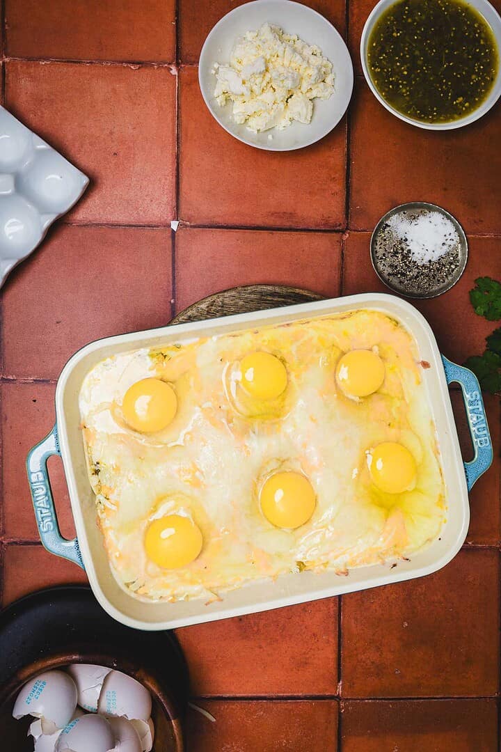 A casserole dish with six cracked eggs on top, surrounded by bowls of cheese, seasoning, and sauce&mdash;perfect for creating a flavorful huevos rancheros breakfast casserole.