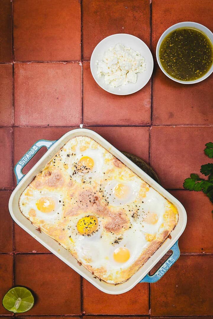 Baked eggs in a casserole dish, reminiscent of a huevos rancheros breakfast casserole, with bowls of cheese and green sauce, on a terracotta tile surface.