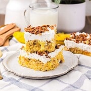 Two dessert bars inspired by Hummingbird Poke Cake, topped with whipped topping and pecans, rest on a plate, with milk and plants in the background.