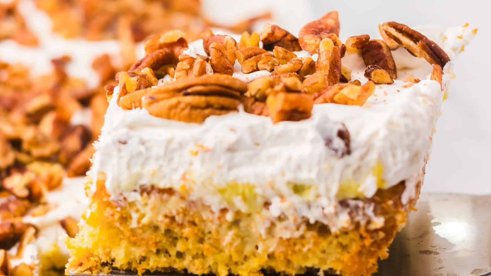 A slice of cake topped with whipped cream and chopped pecans on a metal spatula.