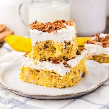 Two frosted cake bars topped with chopped nuts on a plate, reminiscent of classic Hummingbird Poke Cake, with milk and a banana in the background.