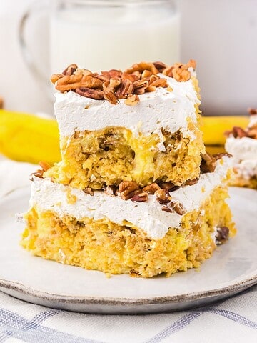 Two frosted cake bars topped with chopped nuts on a plate, reminiscent of classic Hummingbird Poke Cake, with milk and a banana in the background.
