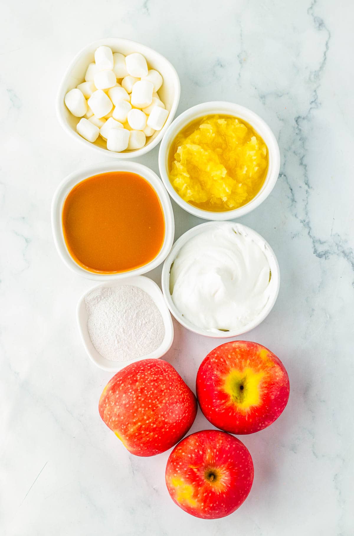 Three red apples, marshmallows, caramel, crushed pineapple, whipped topping, and white powder in bowls come together to create a delicious caramel apple fluff.