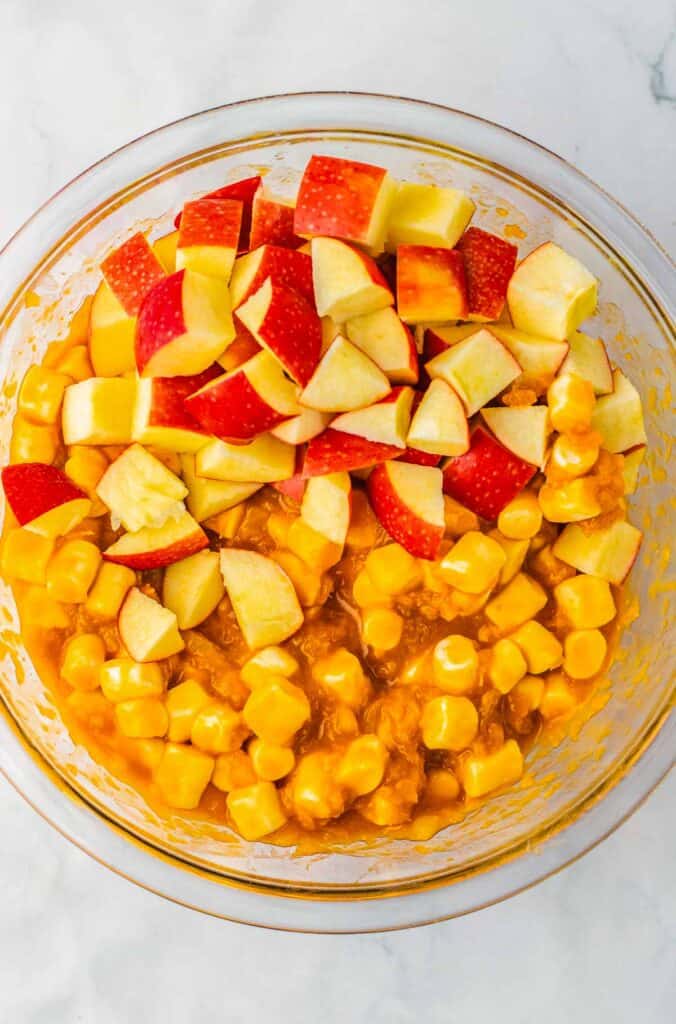 A glass bowl with diced red apples, mini marshmallows, and an orange creamy mixture—this delightful caramel apple fluff is served on a white surface.
