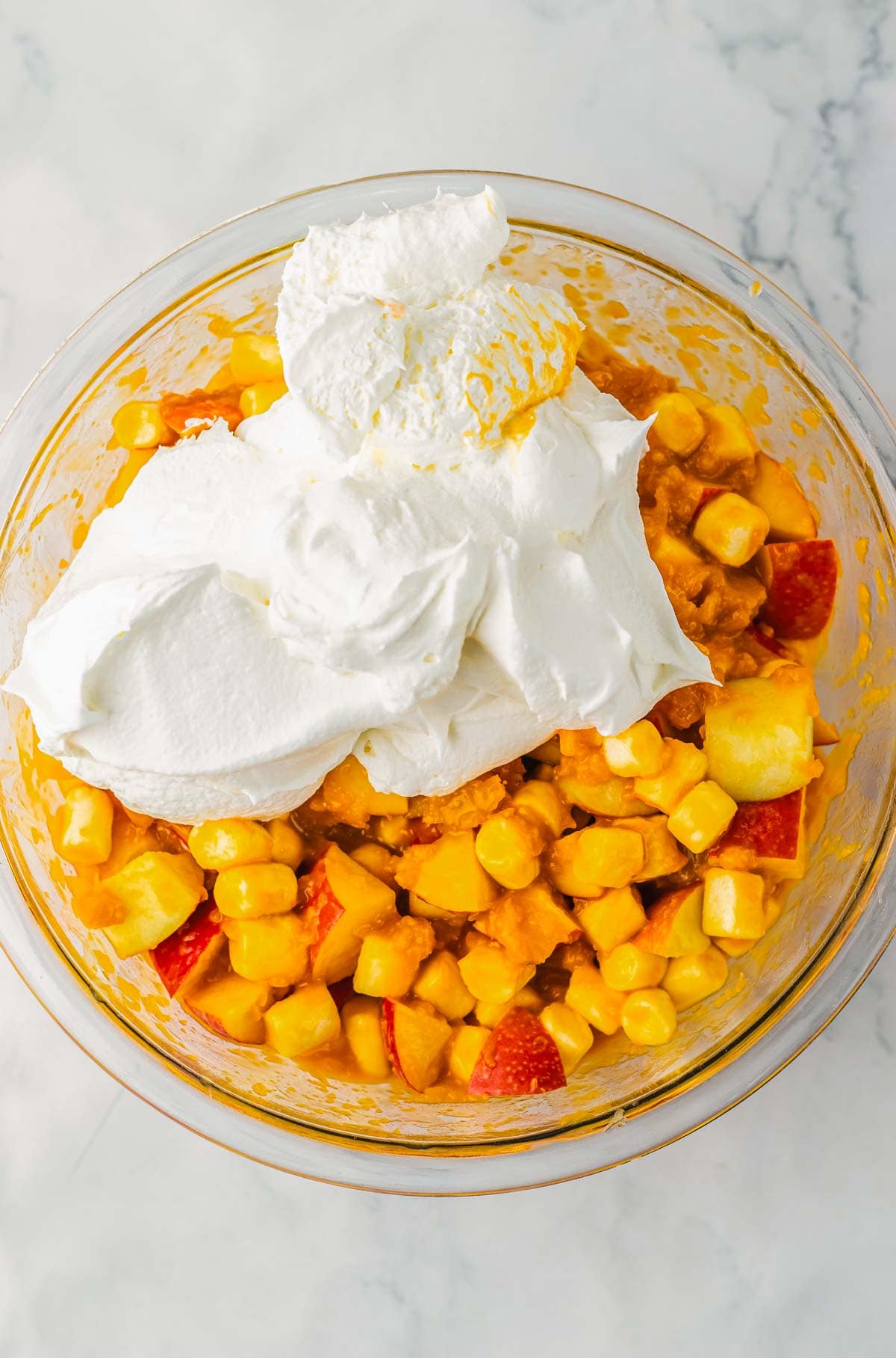 A glass bowl with fruit salad and whipped cream on top, reminiscent of caramel apple fluff, sits on a marble countertop.