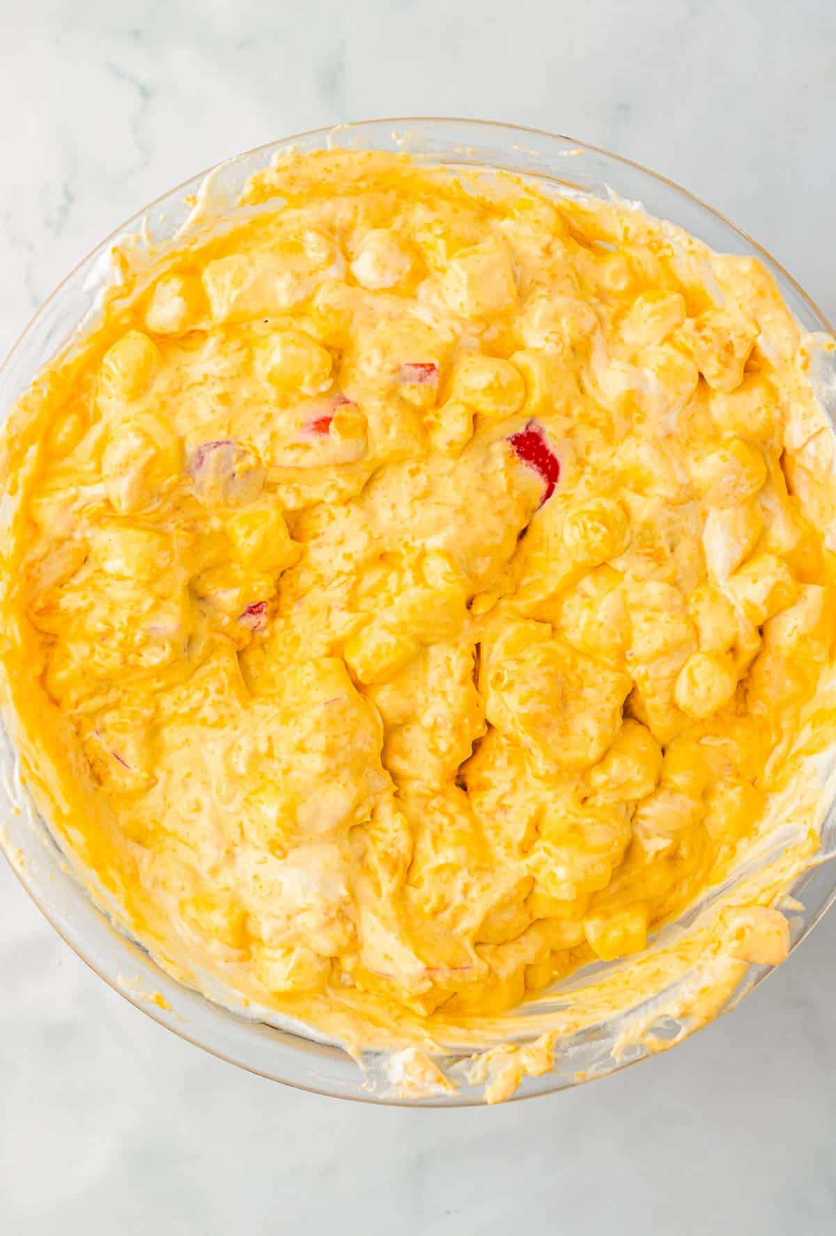 Creamy caramel apple fluff yellow fruit salad with marshmallows and fruit pieces in a glass bowl on a white surface.