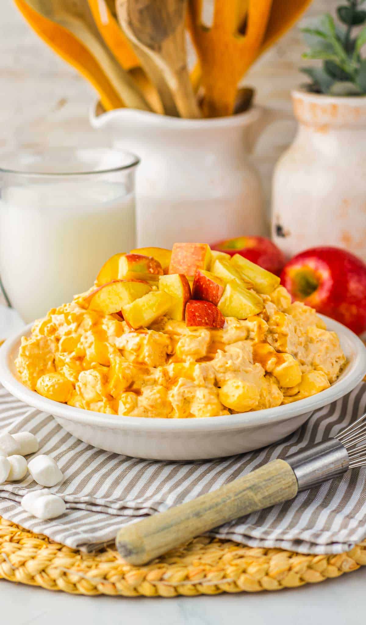 A bowl of creamy caramel apple fluff fruit salad with marshmallows, topped with fresh apple chunks, sits beside a glass of milk.