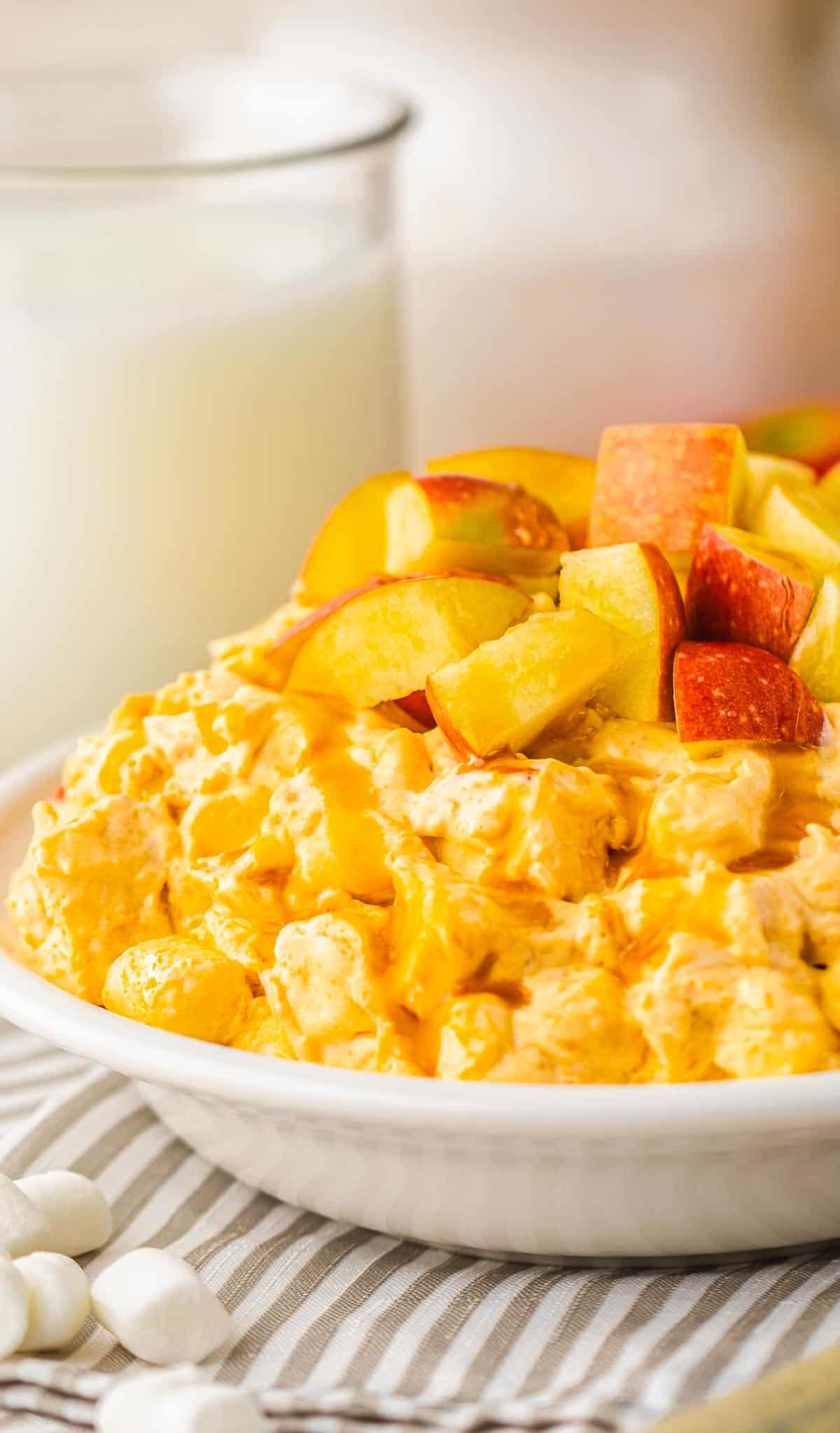 A bowl of creamy fruit salad topped with apple and peach chunks, reminiscent of caramel apple fluff, sits next to a glass of milk.