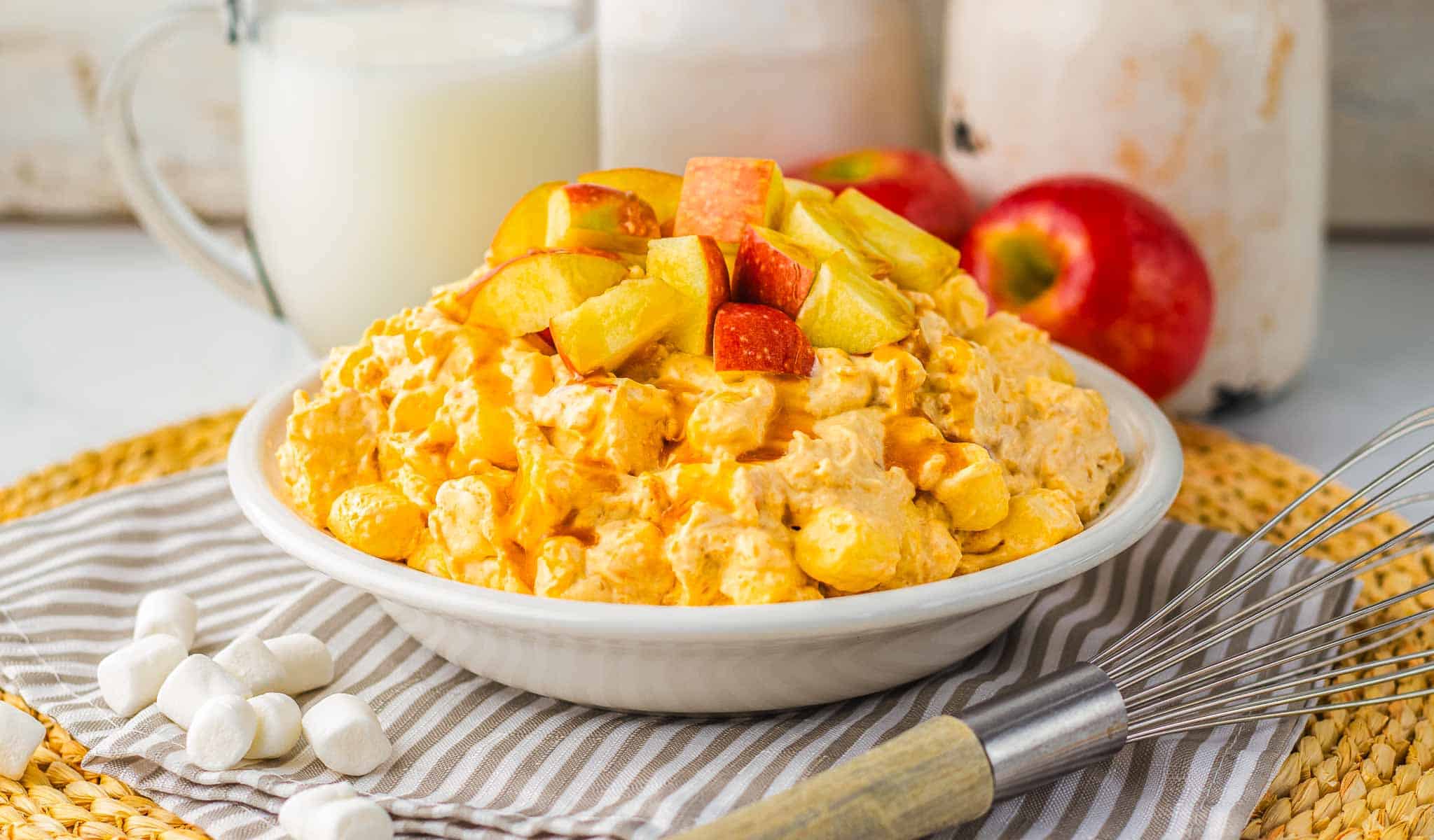 A bowl of creamy fruit salad topped with apple chunks, reminiscent of a classic caramel apple fluff, with marshmallows, apples, and milk in the background.