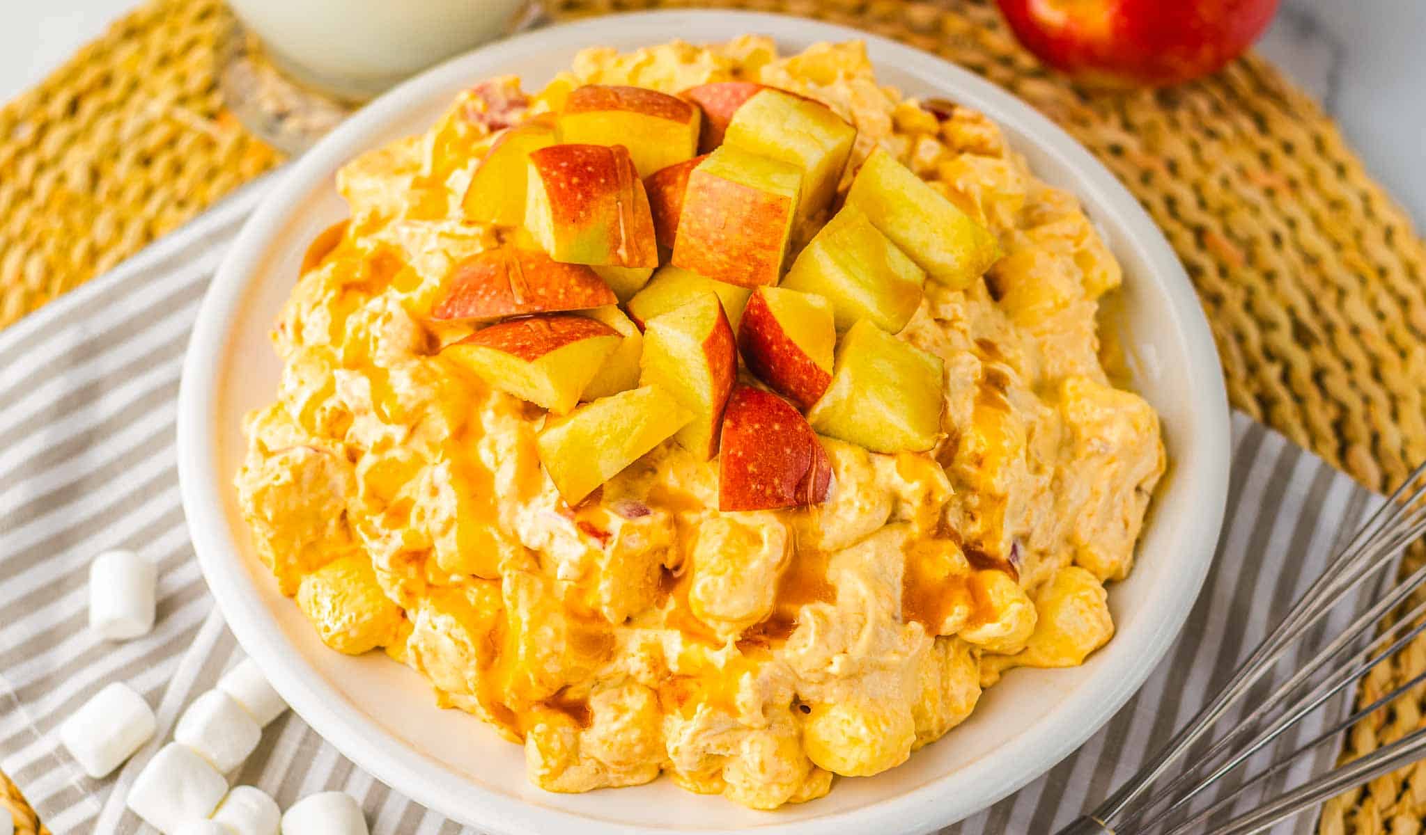 A bowl of creamy caramel apple fluff salad topped with chopped apples, surrounded by marshmallows and a whisk.