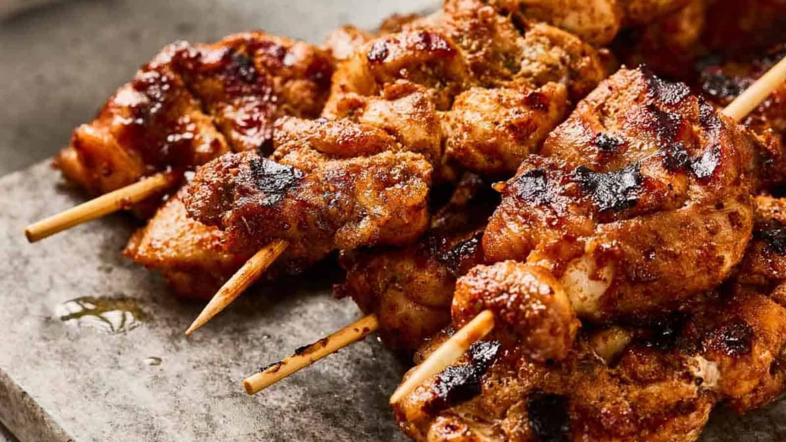 Grilled chicken skewers with spices on a cutting board, with sauce and bread nearby.