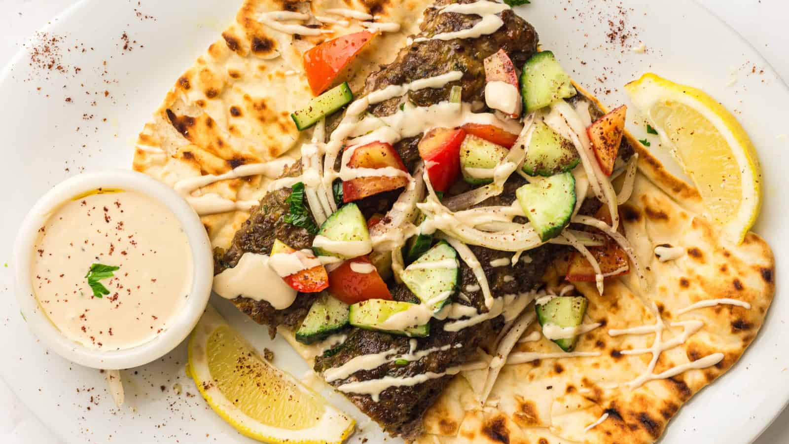 Flatbread topped with grilled meat, diced tomatoes, cucumbers, onions, sauce, and lemon wedges on the side.