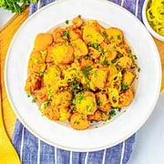 A white plate of seasoned Sheet Pan Mississippi Chicken and Potatoes garnished with herbs sits on a blue cloth next to fresh greens and a drink.