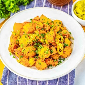A white plate of seasoned potatoes garnished with herbs, inspired by Sheet Pan Mississippi Chicken and Potatoes, sits on a blue-striped cloth, with peppers on the side.