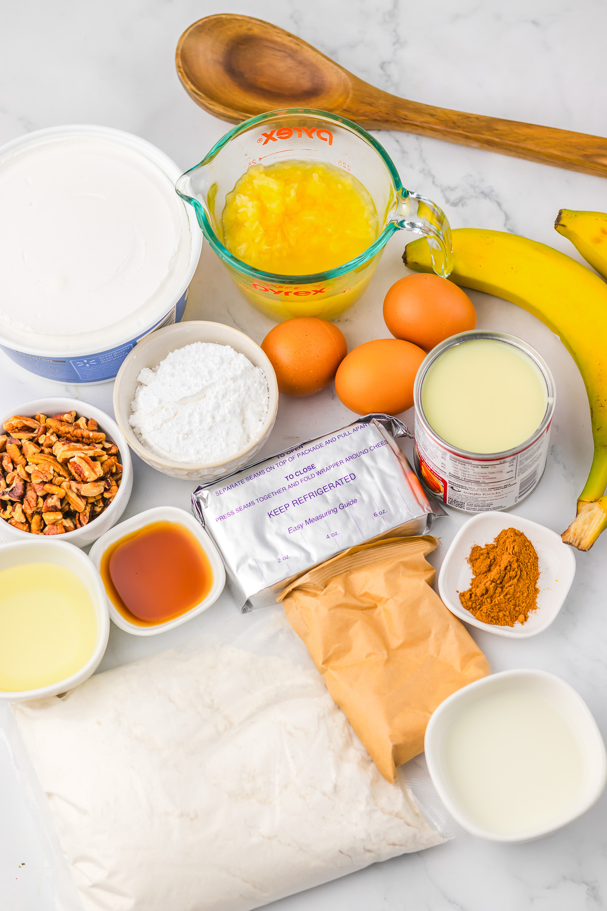Baking ingredients for Hummingbird Poke Cake&mdash;including eggs, bananas, butter, milk, pecans, flour, spices, and vanilla&mdash;arranged on a marble surface.