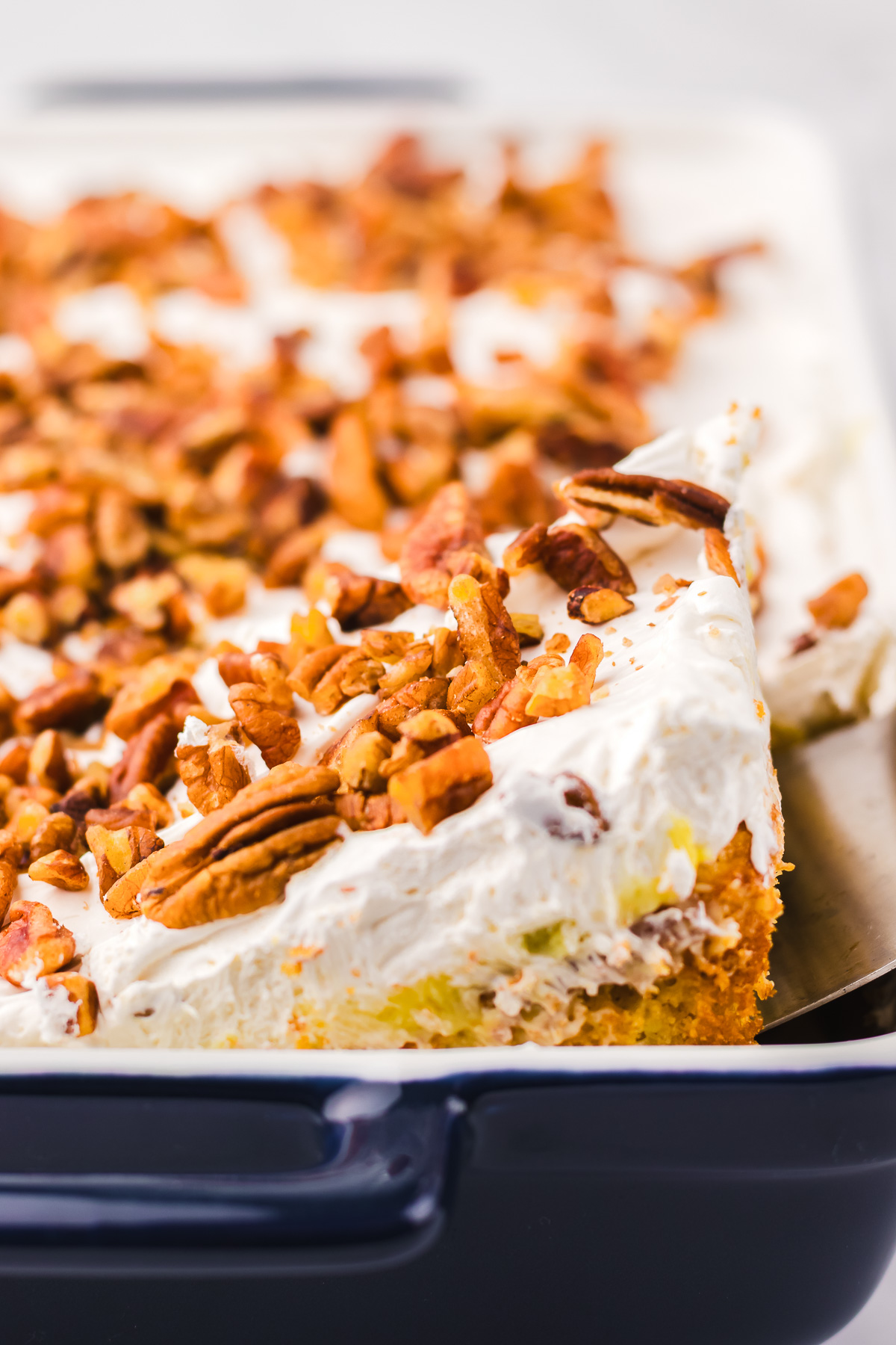A delicious Hummingbird Poke Cake with whipped topping and chopped pecans in a baking dish, with a slice being served.