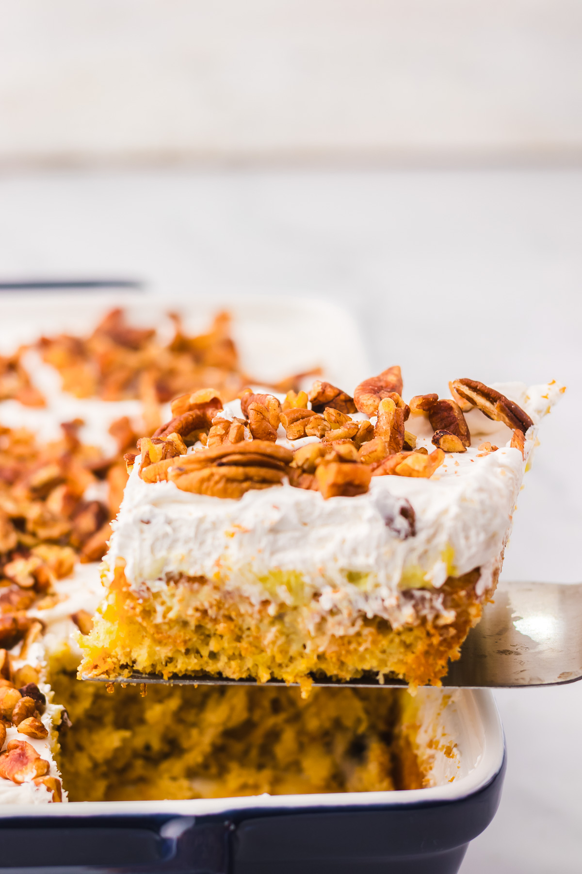 A spatula lifts a slice of Hummingbird Poke Cake, topped with whipped topping and chopped pecans, from a baking dish.