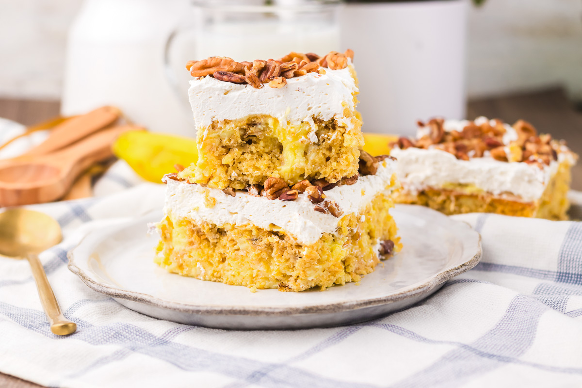 Two squares of Hummingbird Poke Cake with whipped topping and pecans, stacked on a plate, with a third piece beside.