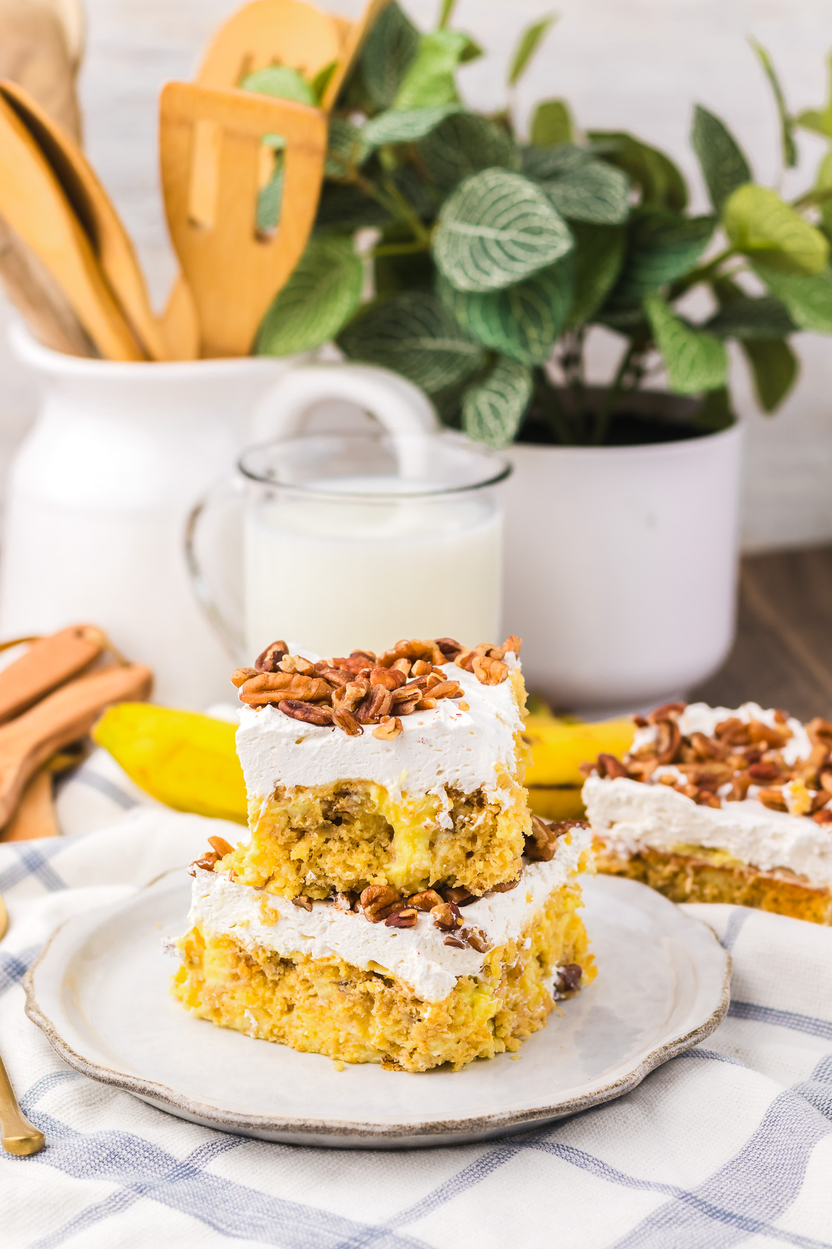 Two frosted dessert bars, inspired by Hummingbird Poke Cake, with nuts on a plate, a glass of milk, and a plant in the background.