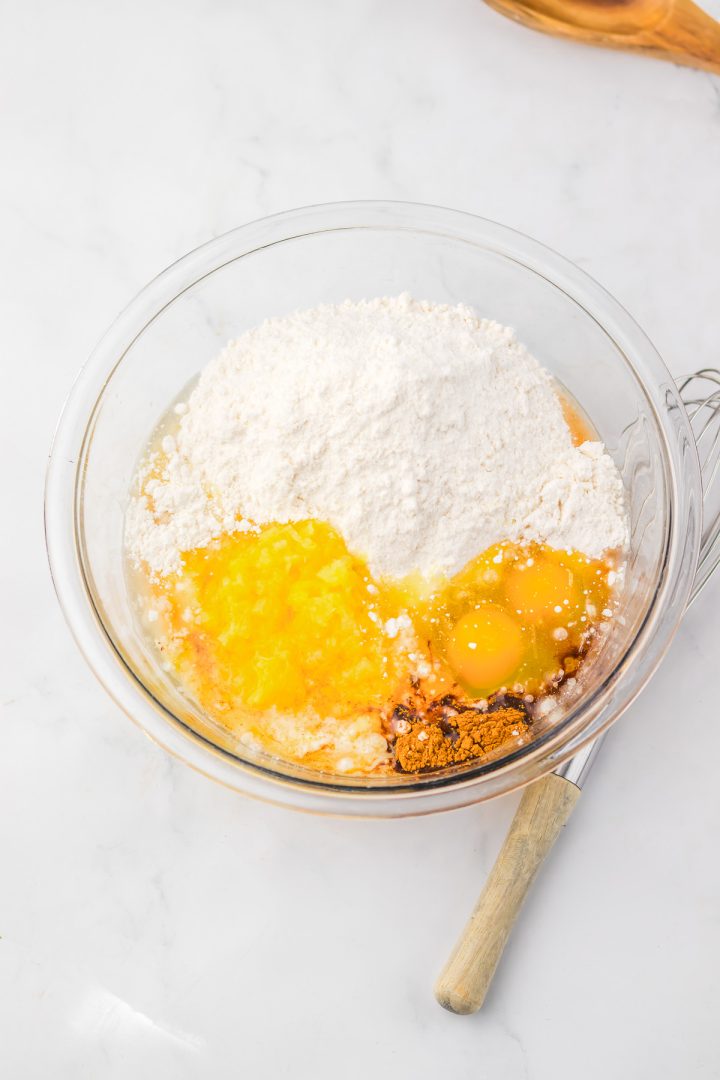 A glass bowl with flour, eggs, spices, and wet ingredients, ready to be mixed for a delicious Hummingbird Poke Cake, sits on a marble surface.