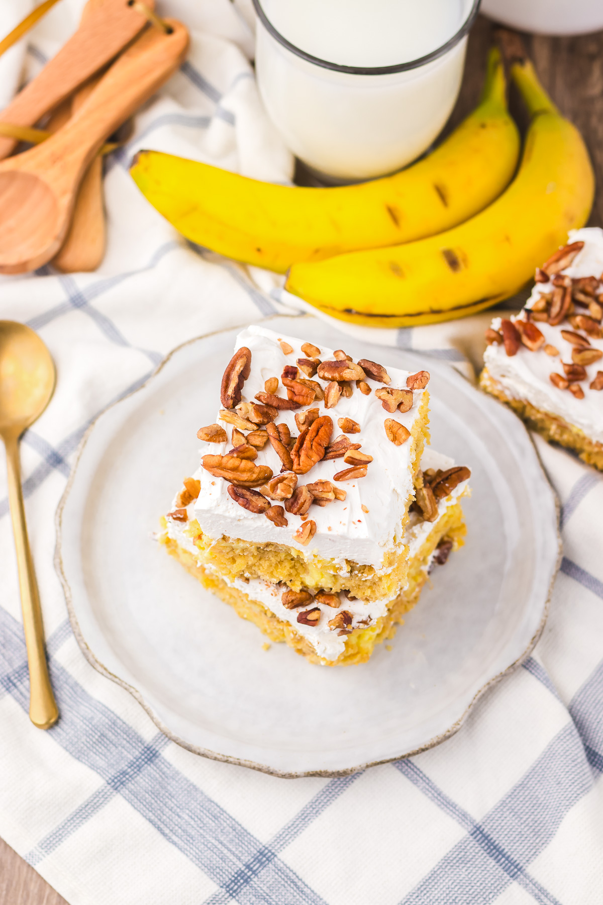 Two slices of frosted Hummingbird Poke Cake with pecans rest on a plate, accompanied by fresh bananas and a glass of milk nearby.