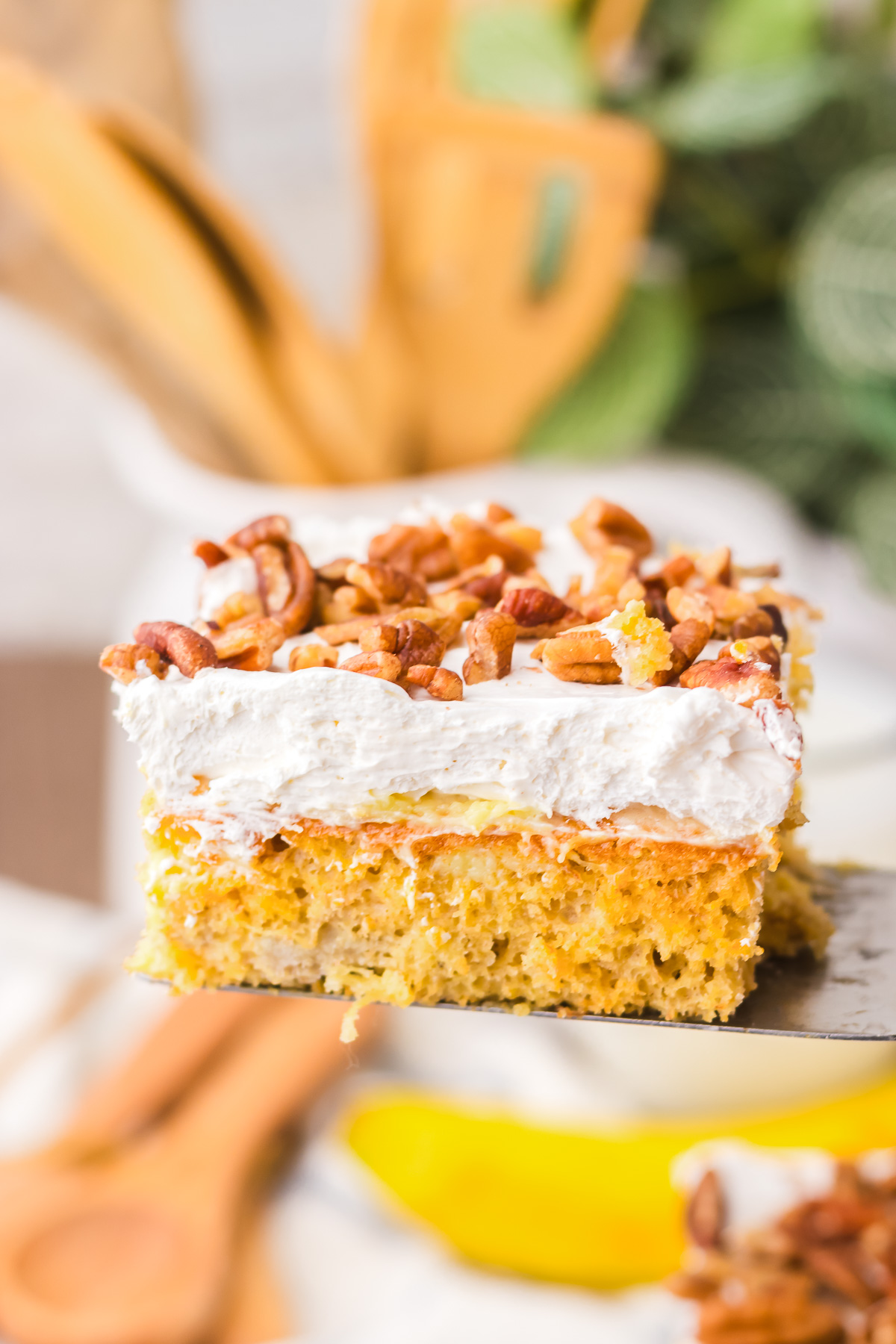A close-up of a Hummingbird Poke Cake slice topped with whipped cream and chopped pecans, held above a blurred background.