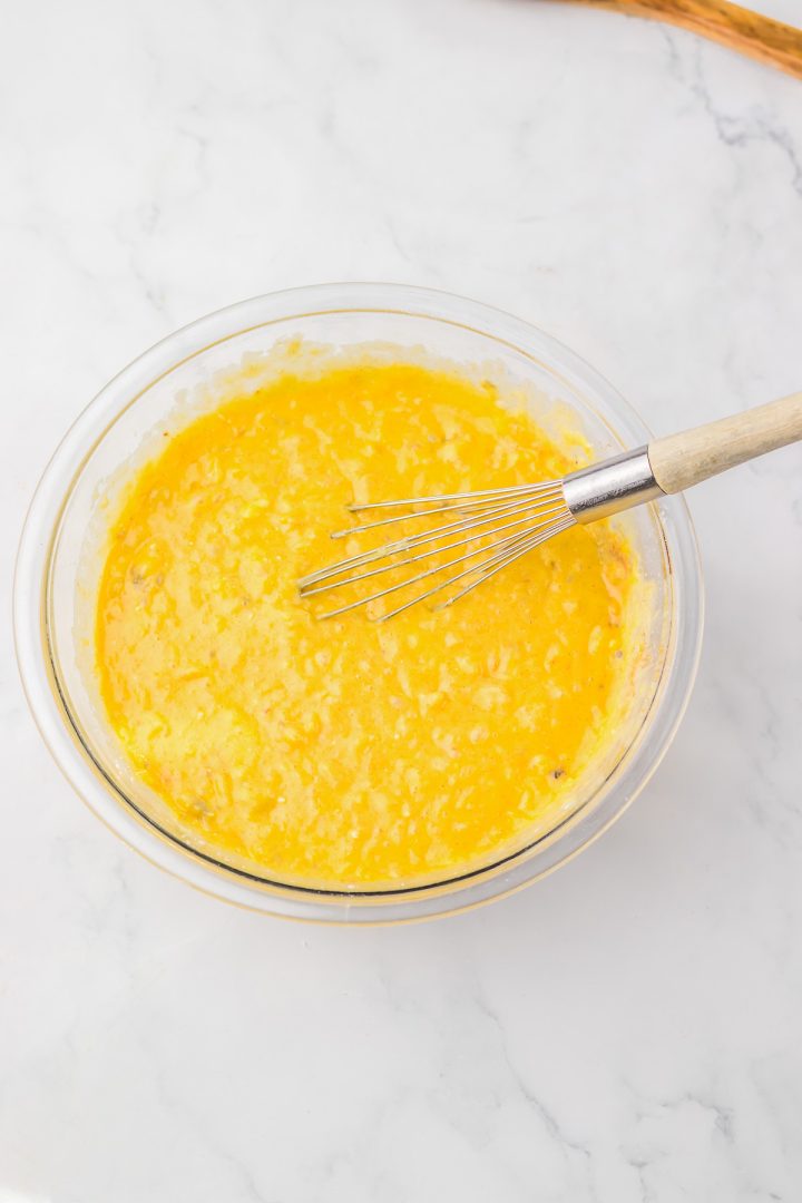 A whisk in a glass bowl of yellow, slightly lumpy Hummingbird Poke Cake batter rests on a white marble surface.