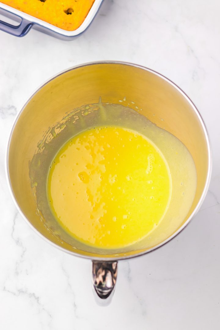 A metal mixing bowl with a yellow liquid batter inside, perfect for making Hummingbird Poke Cake, sits on a white marble surface.