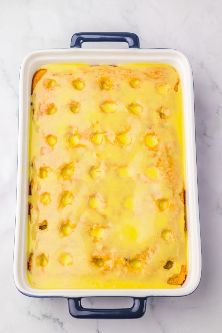 A rectangular Hummingbird Poke Cake with holes, covered in yellow glaze, sits in a white baking dish with blue handles.