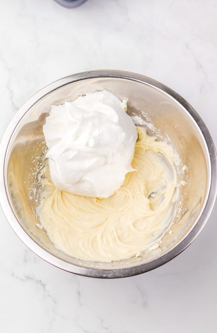 A metal bowl with whipped cream and cream cheese mixture, perfect for topping your Hummingbird Poke Cake, sits on a white marble surface.