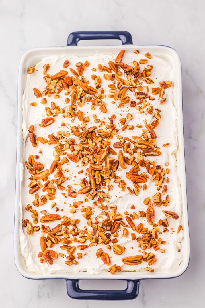 Rectangular Hummingbird Poke Cake topped with white frosting and chopped pecans in a blue-handled baking dish.