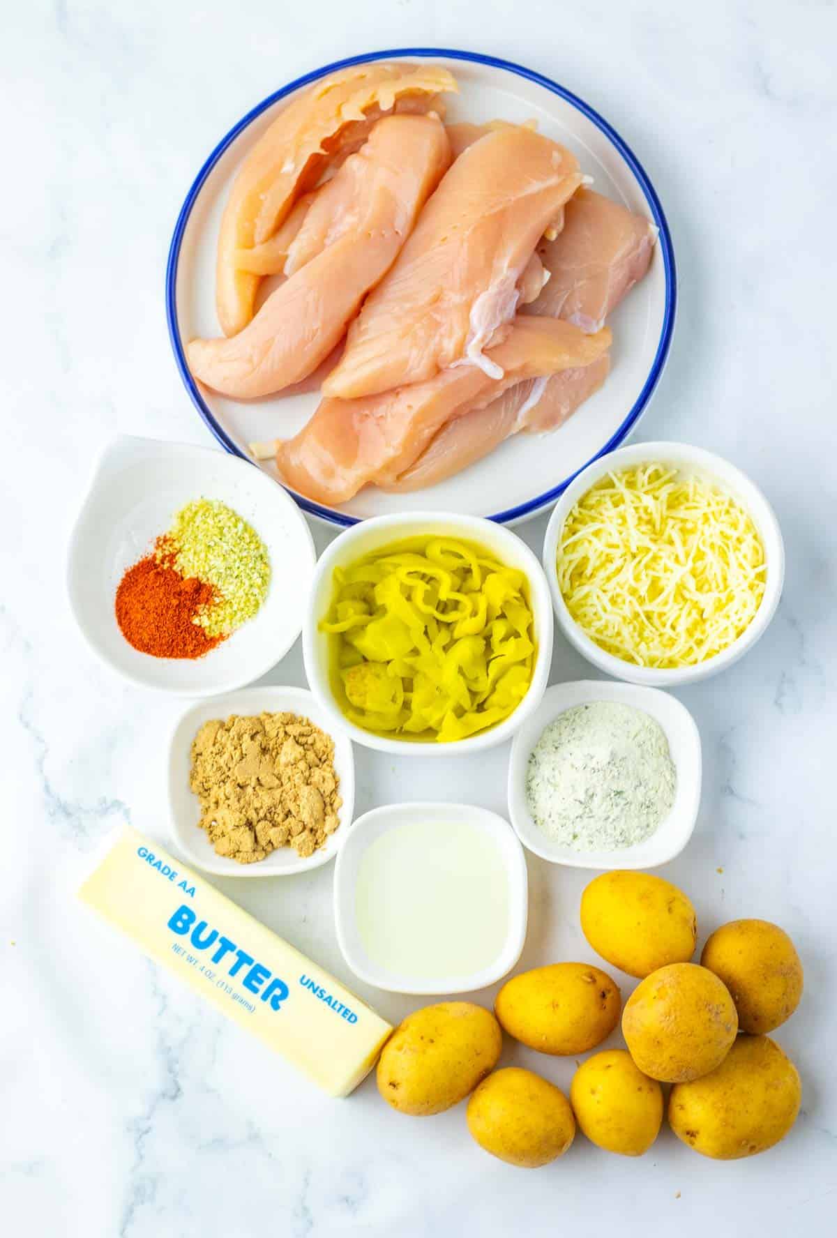 Ingredients for Sheet Pan Mississippi Chicken and Potatoes&mdash;raw chicken breasts, shredded cheese, potatoes, butter, seasonings, peppers, and milk&mdash;arranged on a marble surface.