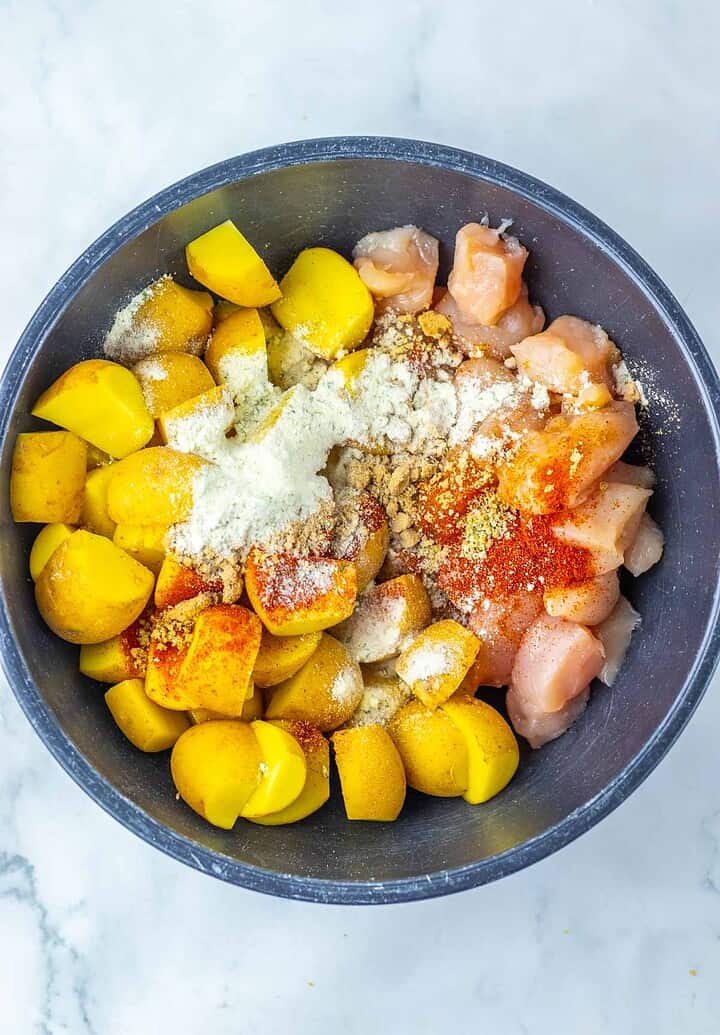 A bowl with diced chicken, yellow potatoes, flour, and spices on a white marble surface&mdash;perfect ingredients for Sheet Pan Mississippi Chicken and Potatoes.