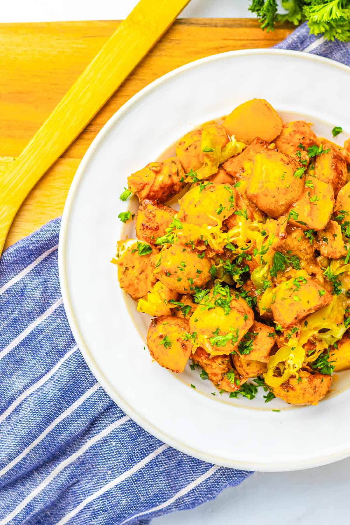 White plate of cooked sausage and potatoes garnished with herbs, on a blue striped cloth with a wooden spoon&mdash;reminiscent of a comforting Sheet Pan Mississippi Chicken and Potatoes dinner.
