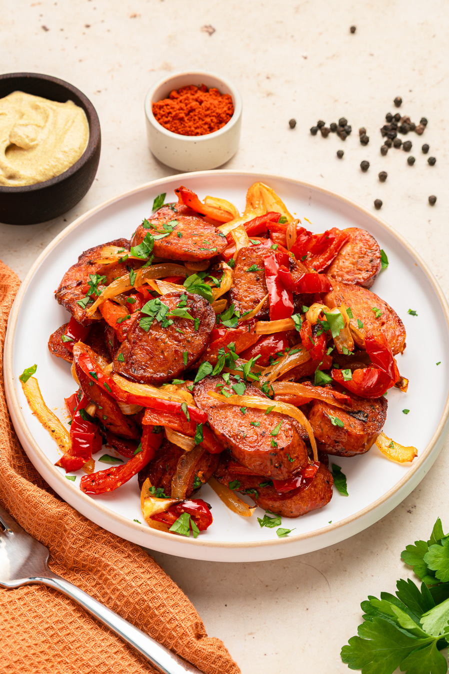 Air fryer kielbasa saut&eacute;ed with onions, red peppers, and herbs on a white plate, with spices and mustard nearby.
