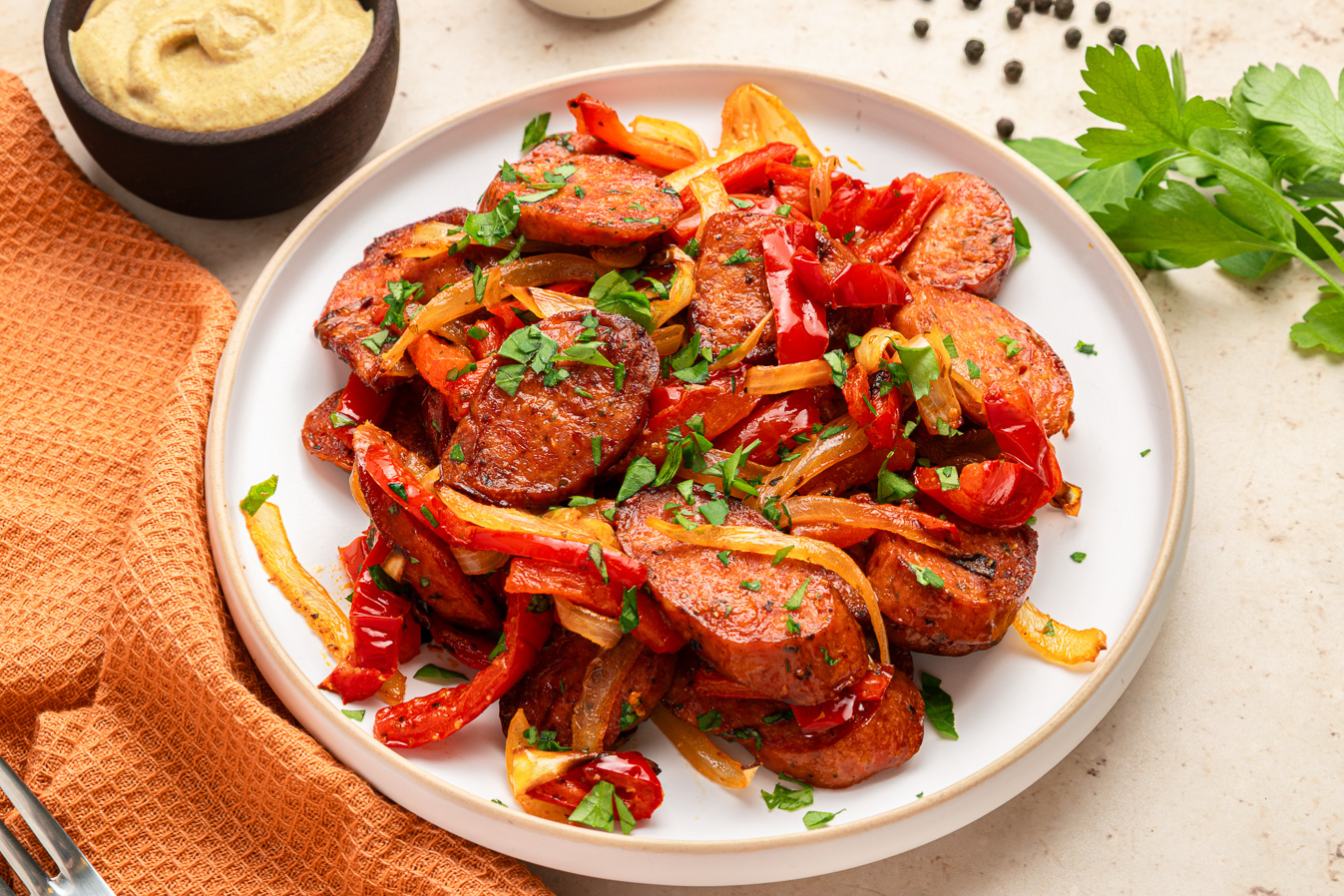 Saut&eacute;ed air fryer kielbasa, red peppers, and onions garnished with parsley on a white plate with a napkin nearby.