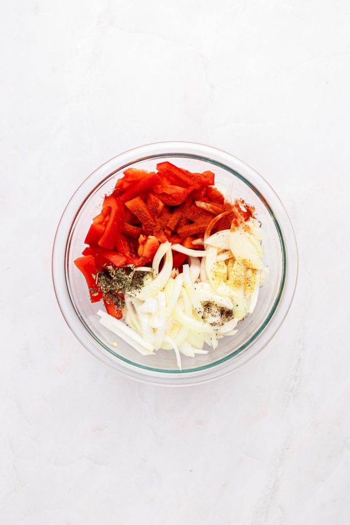 Glass bowl with sliced onions, red bell peppers, and various spices on a white marble surface&mdash;perfect ingredients to pair with air fryer kielbasa for a flavorful and easy meal.