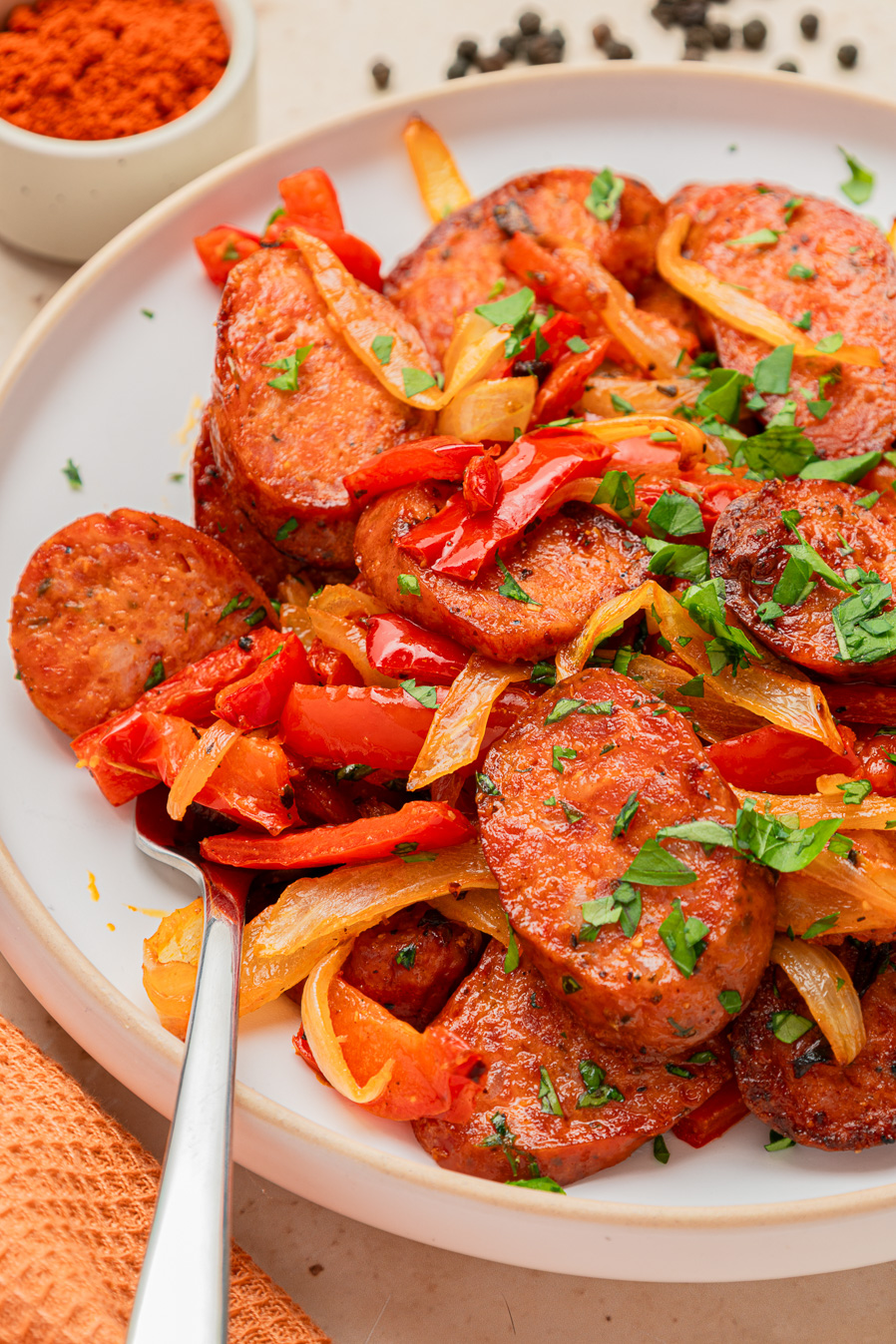 Air fryer kielbasa sliced with saut&eacute;ed onions, red peppers, and parsley is served on a white plate with a fork.