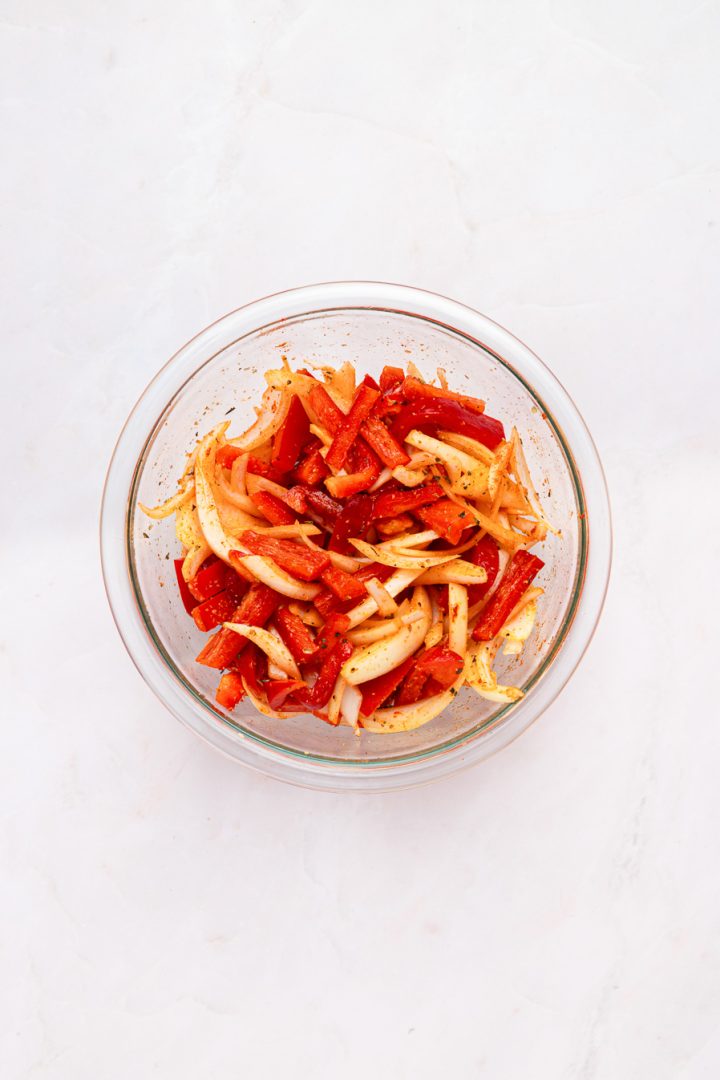 A glass bowl filled with sliced red bell peppers and onions mixed with seasoning sits on a white surface, ready to be paired with air fryer kielbasa for a quick and flavorful meal.