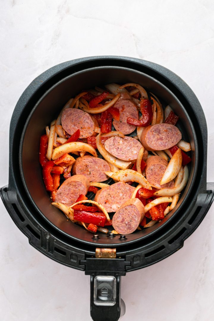 Sliced air fryer kielbasa, red bell peppers, and onions in an air fryer basket, ready to be cooked.