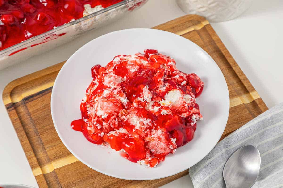 A white plate with vintage Cherries in the Snow Dessert, topped with whipped topping and cake, sits on a wooden board next to a spoon.