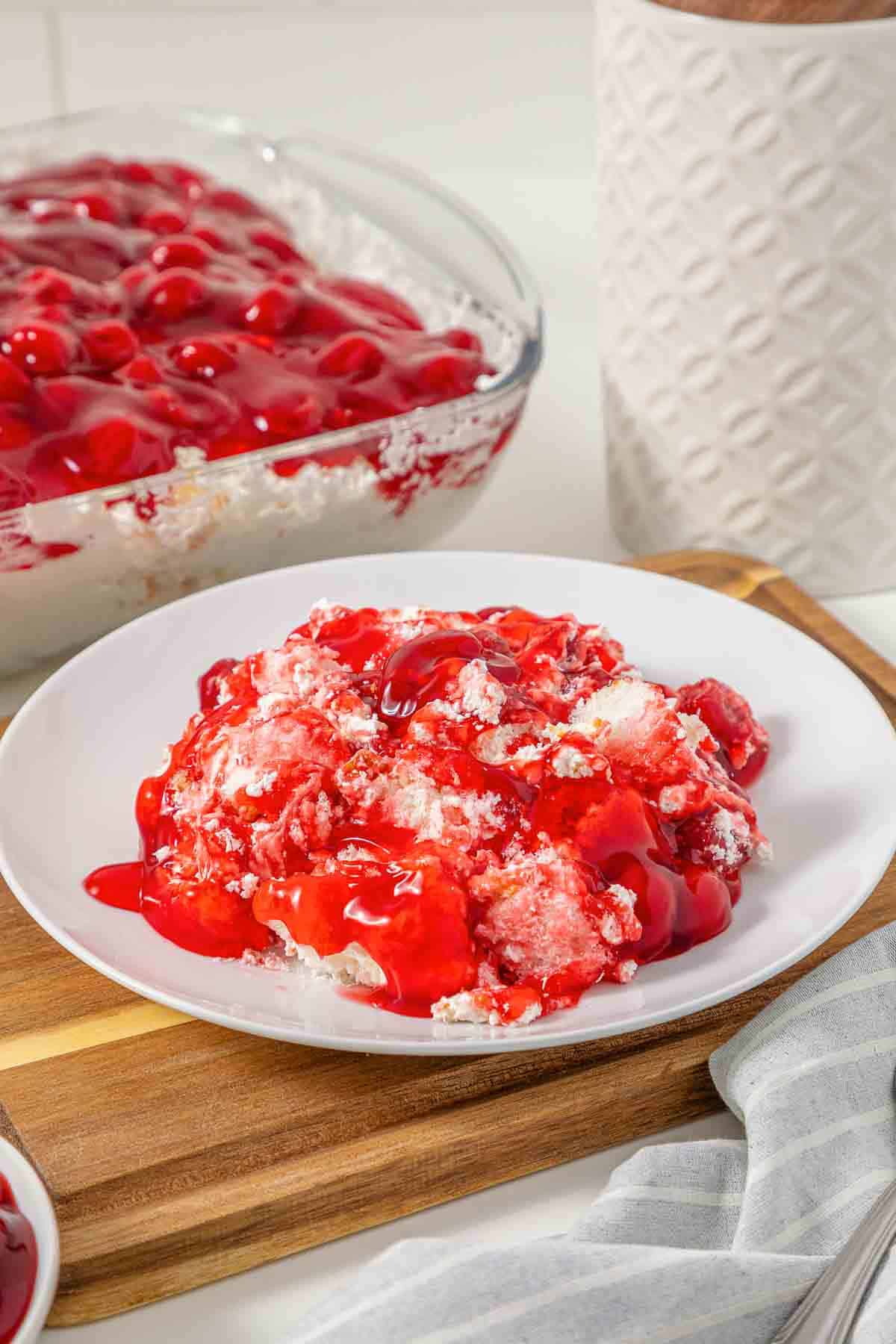 A serving of cherry dump cake on a white plate, reminiscent of the vintage Cherries in the Snow Dessert, with more cake in a glass dish in the background.