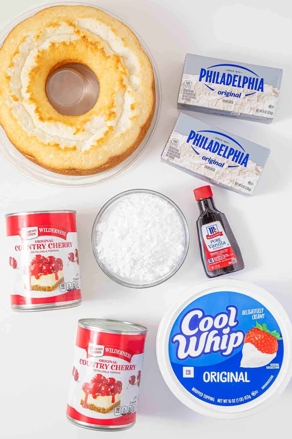 A round cake, two cream cheese blocks, cherry filling cans, powdered sugar, vanilla, and Cool Whip on a table—everything you need to create the vintage Cherries in the Snow Dessert.