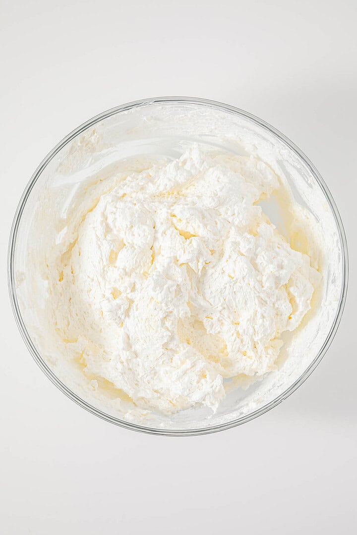 A glass bowl filled with whipped white frosting or cream, reminiscent of the vintage Cherries in the Snow Dessert, viewed from above on a white surface.