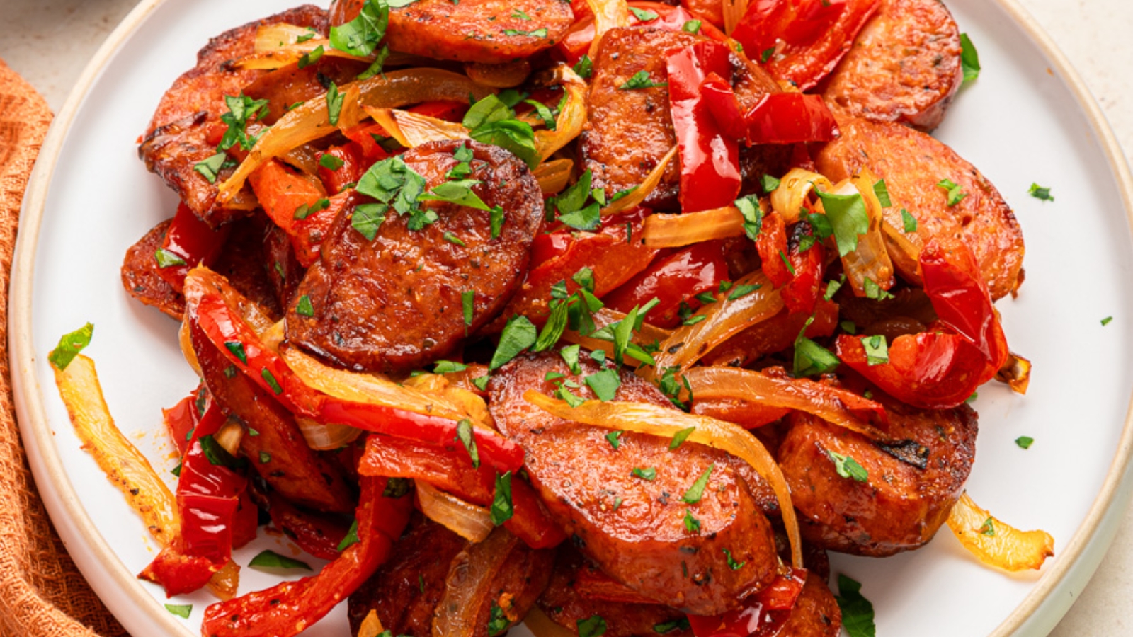 Sliced sausage with sautéed red and yellow peppers and herbs on a white plate, napkin and parsley nearby.